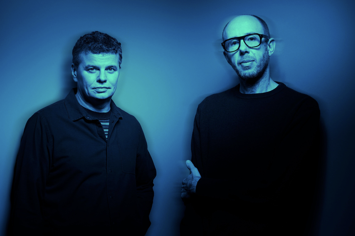 English electronic music duo The Chemical Brothers posing together against a dark blue background with atmospheric lighting